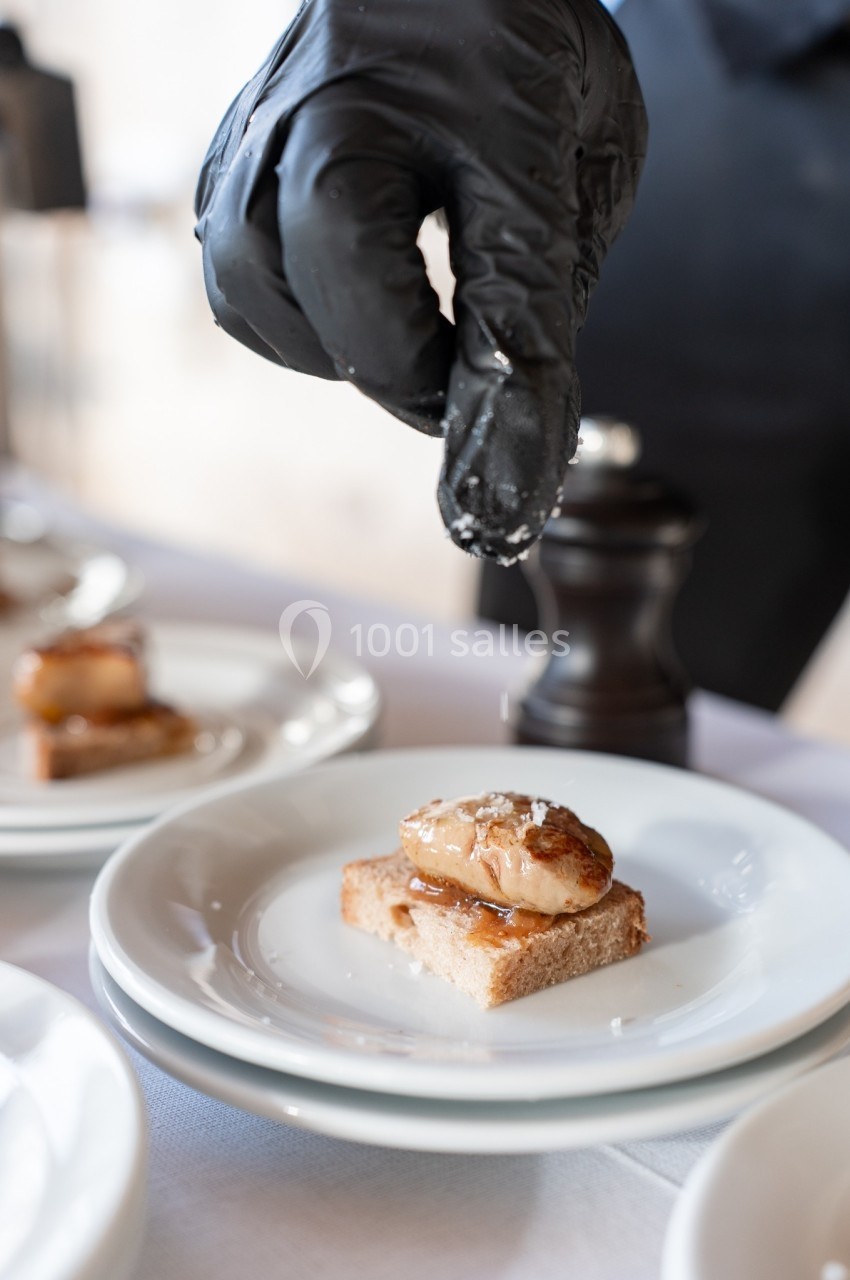 Un chef saupoudre du sel sur un morceau de foie gras posé sur une tranche de pain, présenté dans une assiette blanche.