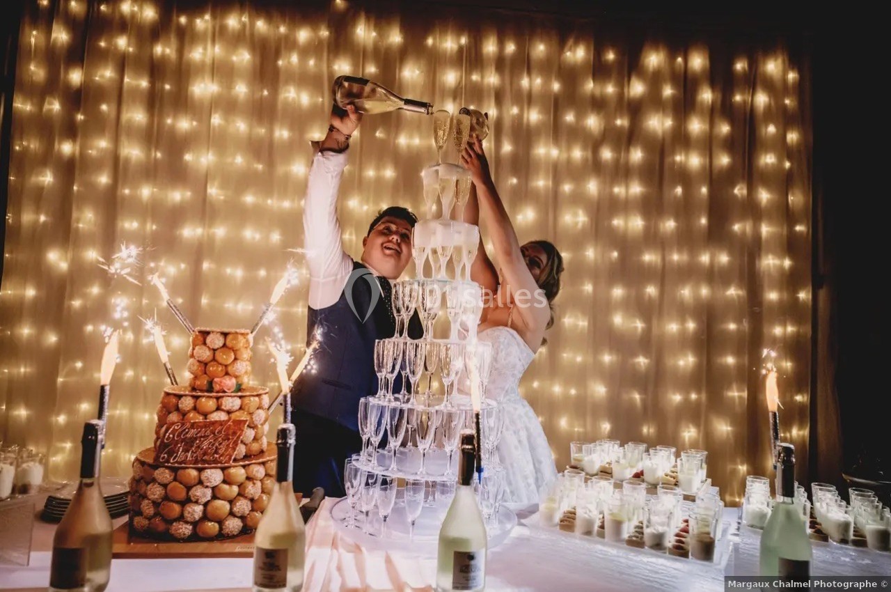 Un couple verse du champagne dans une pyramide de verres lors d'une réception, avec des lumières en arrière-plan.