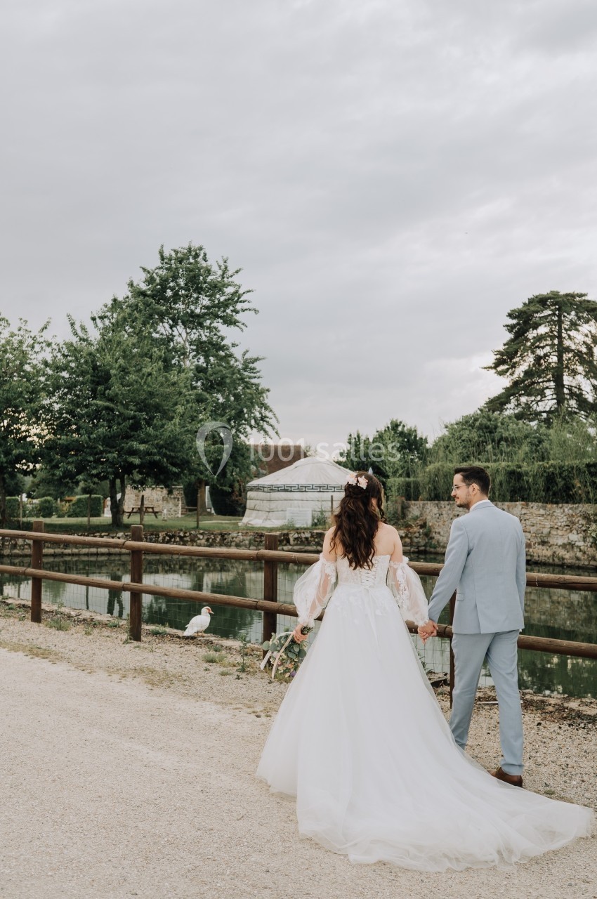 Un couple en tenue de mariage marche main dans la main près d'un étang, entouré de verdure et d'une clôture en bois.