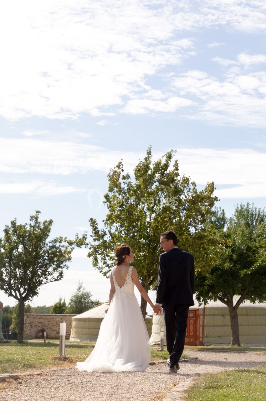Un couple en tenue de mariage marche main dans la main sur un chemin, entouré d'arbres et de yourtes sous un ciel dégagé.