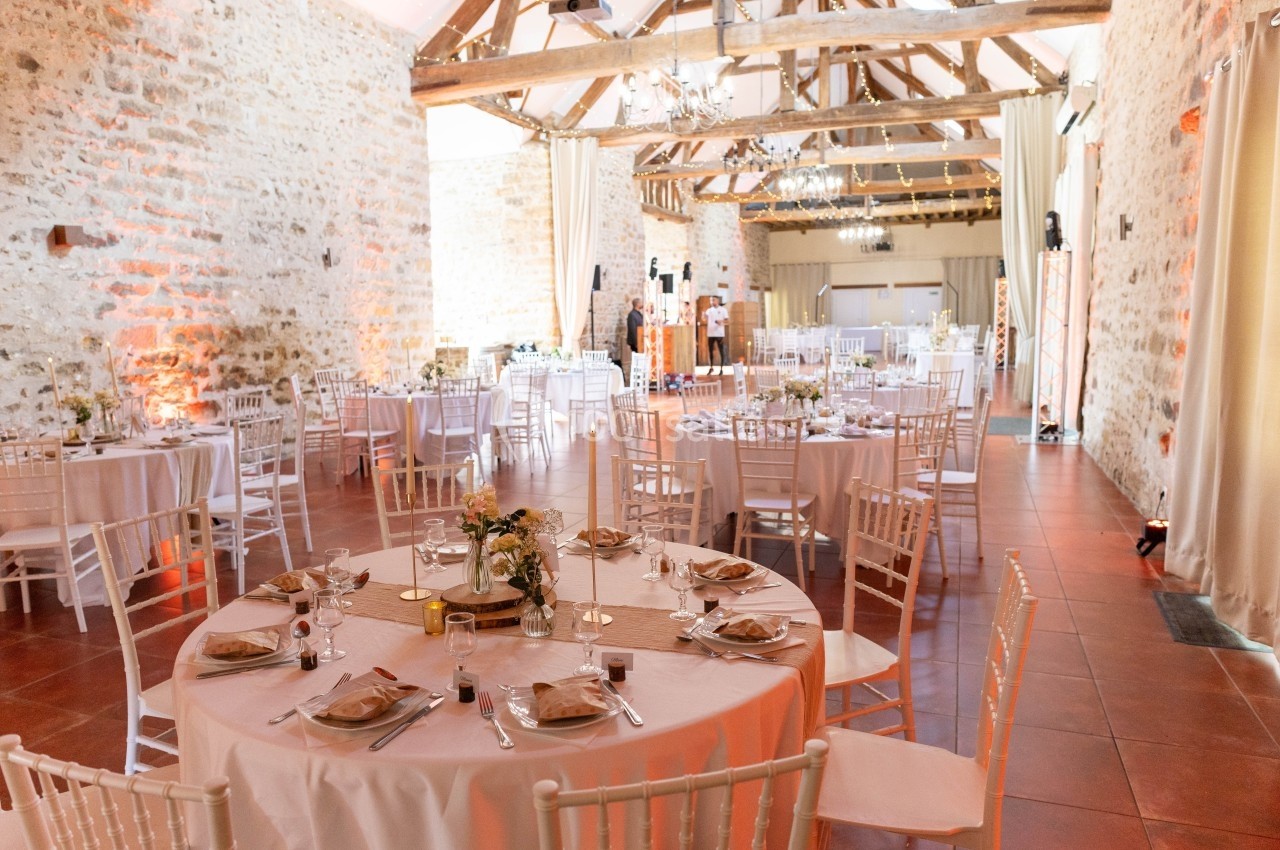Salle de réception lumineuse avec murs en pierre, poutres apparentes et tables rondes décorées pour un événement.