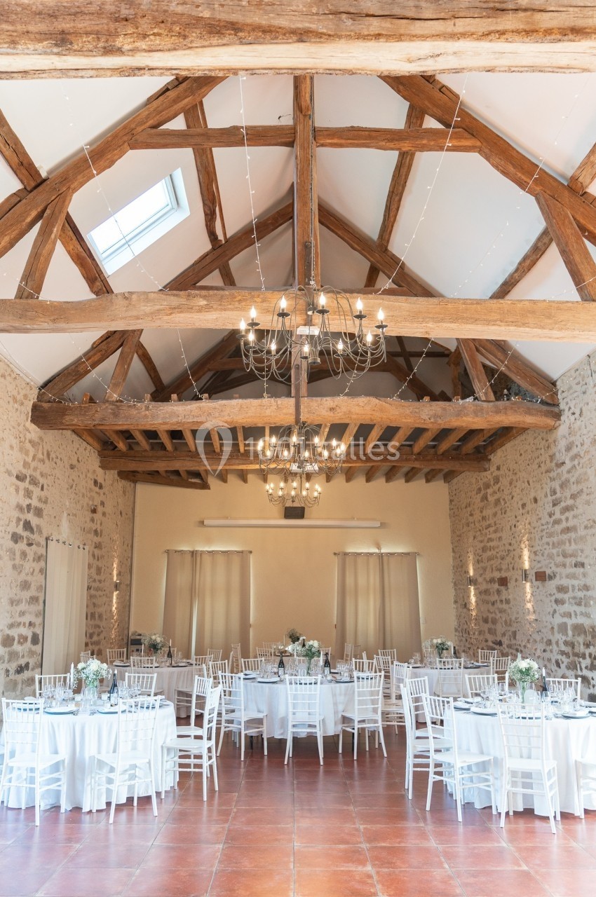 Salle de réception lumineuse avec poutres apparentes, tables rondes dressées et chaises blanches.