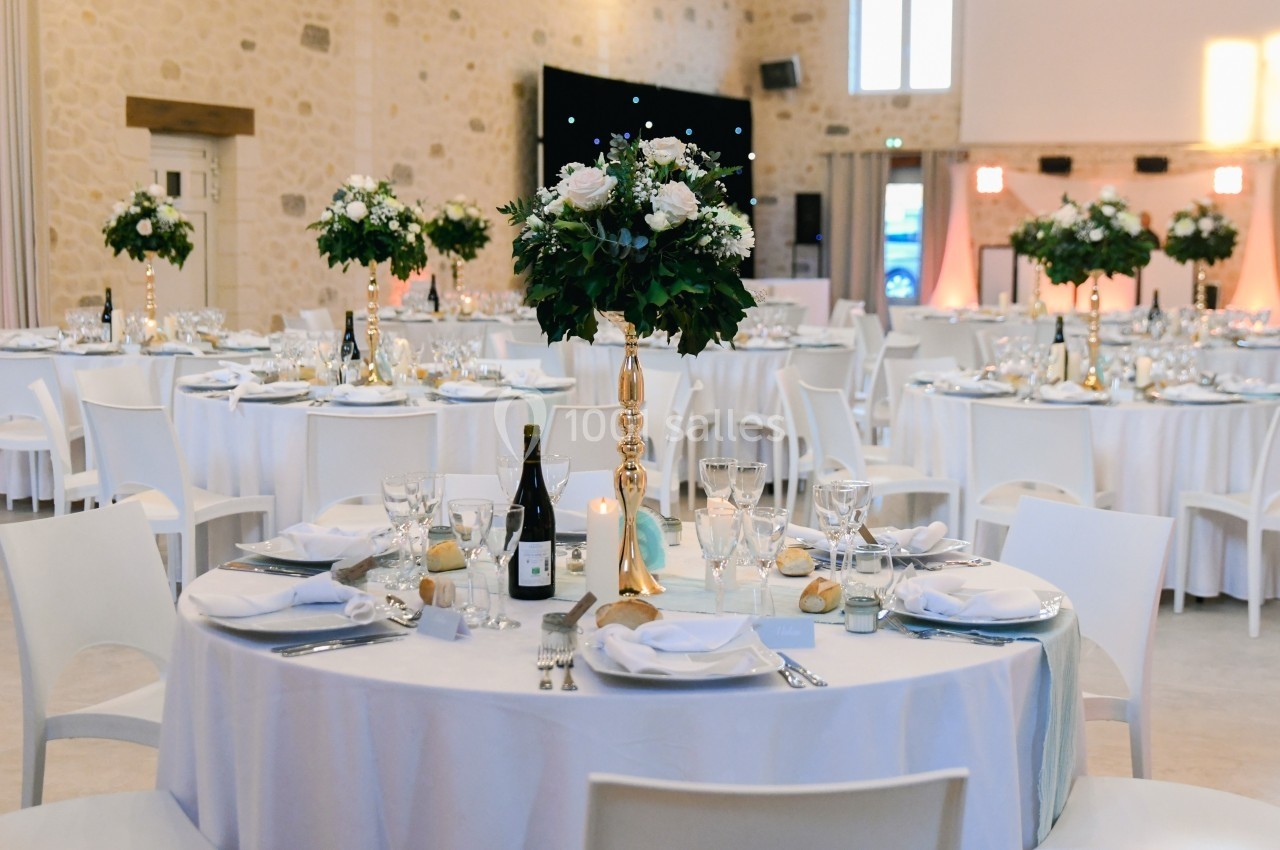 Salle de réception décorée avec des tables rondes, nappes blanches, chandeliers dorés et arrangements floraux.