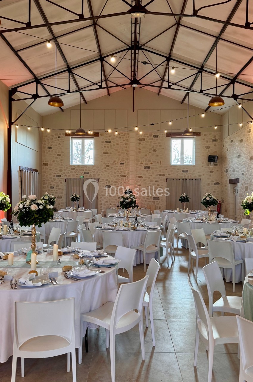 Salle de réception lumineuse avec tables rondes dressées, chaises blanches et décorations florales élégantes.
