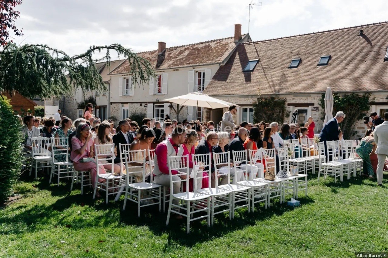 Groupe de personnes assises sur des chaises blanches dans un jardin, devant des maisons en arrière-plan.