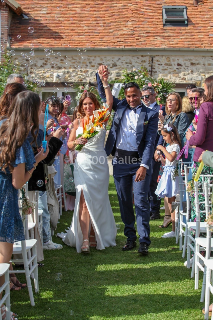 Un couple de mariés marche main dans la main sous les applaudissements des invités dans un jardin ensoleillé.