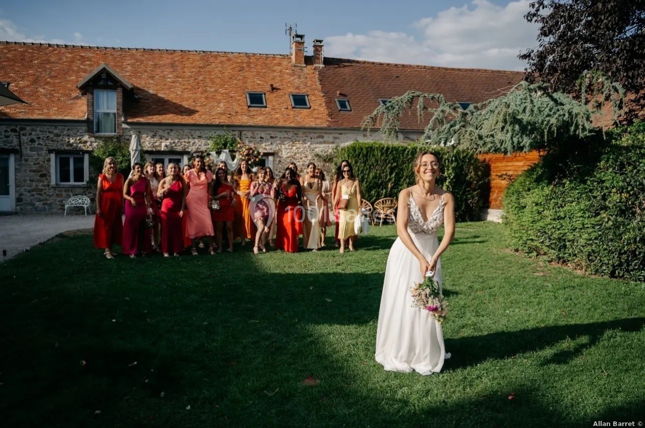 Une mariée en robe blanche s'apprête à lancer son bouquet devant un groupe d'invitées dans un jardin ensoleillé.
