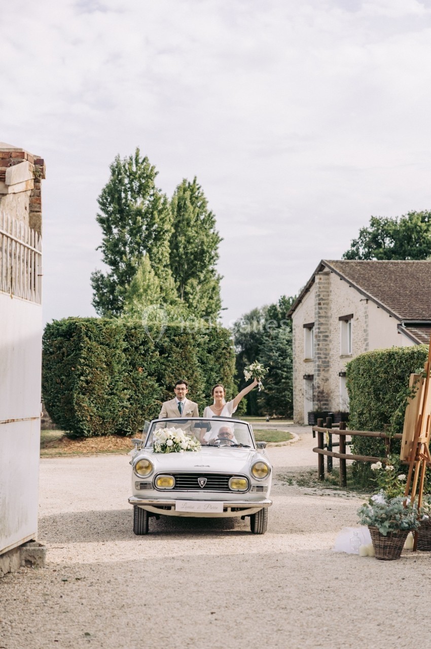 Un couple souriant dans une voiture décapotable ancienne, avançant sur une allée gravillonnée devant des bâtiments ruraux.