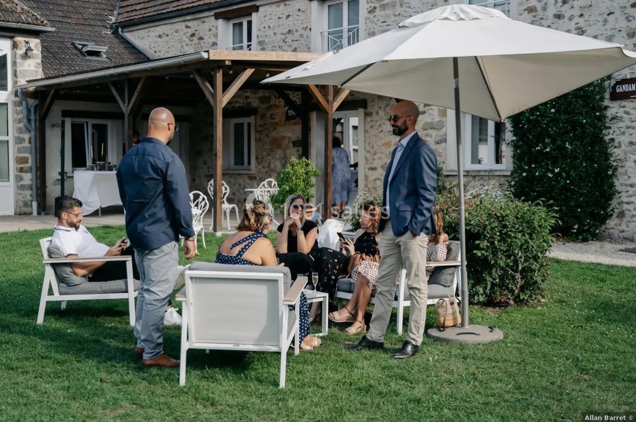 Des personnes discutent en plein air, assises et debout sous un parasol dans le jardin d'une maison en pierre.