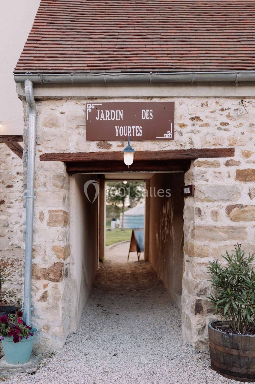 Passage voûté en pierre menant à un jardin, avec une enseigne ’Jardin des yourtes’ au-dessus de l'entrée.
