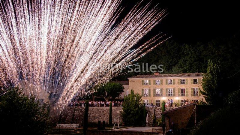 Feu d'artifice illuminant le ciel nocturne devant un grand bâtiment éclairé entouré de végétation.