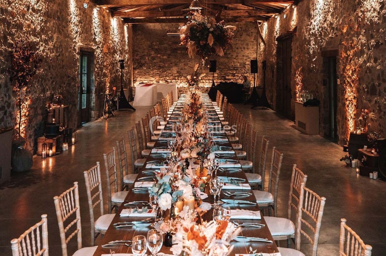 Salle en pierre avec une longue table décorée de fleurs et bougies, éclairée par une lumière chaleureuse.