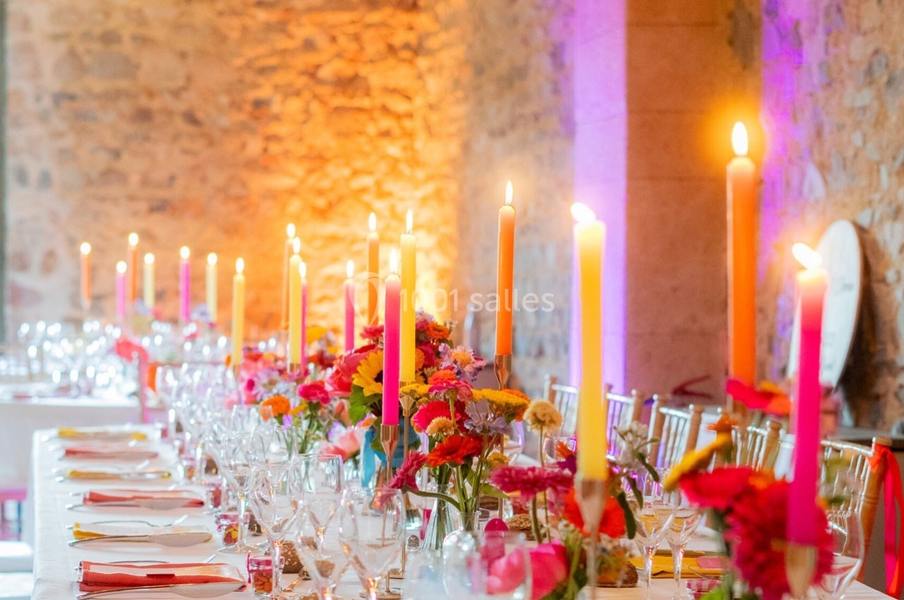 Grande table décorée de bougies allumées, fleurs colorées et vaisselle élégante dans une salle aux murs en pierre.
