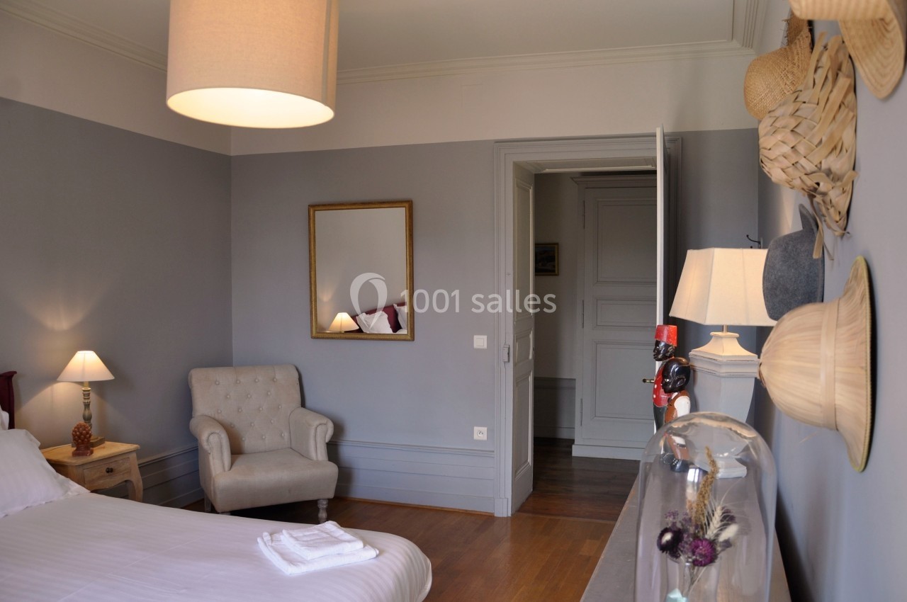 Chambre lumineuse avec murs gris, lit, fauteuil beige, miroir doré, lampes allumées et décoration murale en chapeaux.