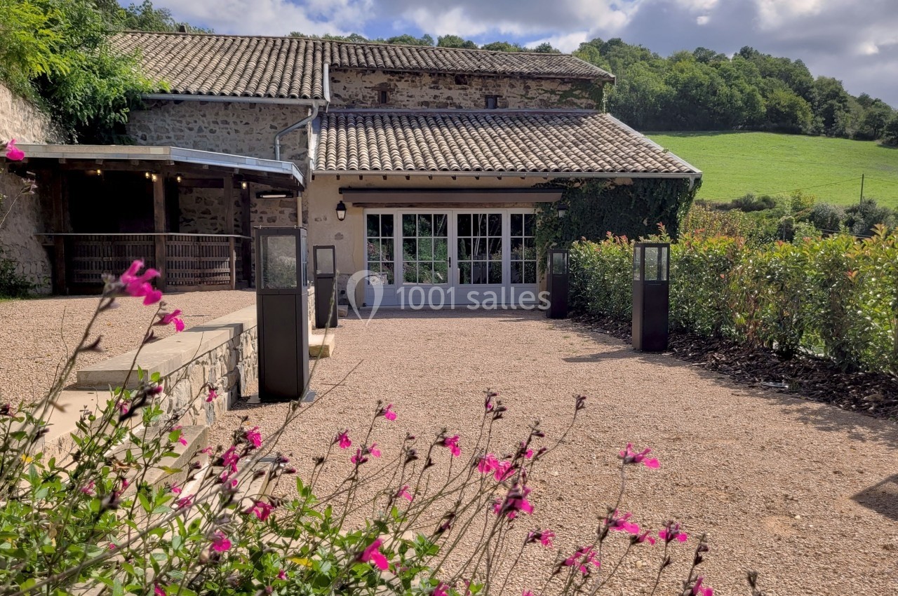 Cour d'une maison en pierre avec terrasse, fleurs roses au premier plan et colline verdoyante en arrière-plan.