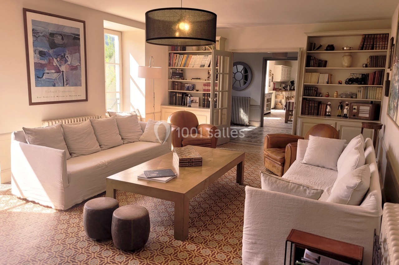 Salon lumineux avec canapés blancs, table basse en bois, étagères remplies de livres et grande fenêtre laissant entrer la…