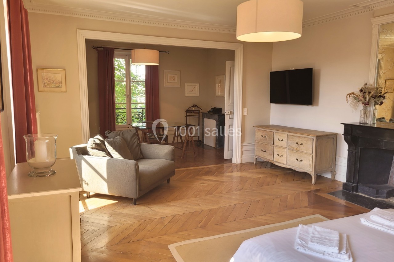 Salon lumineux avec parquet en bois, canapé, bureau, cheminée, télévision murale et grandes fenêtres avec rideaux rouges.