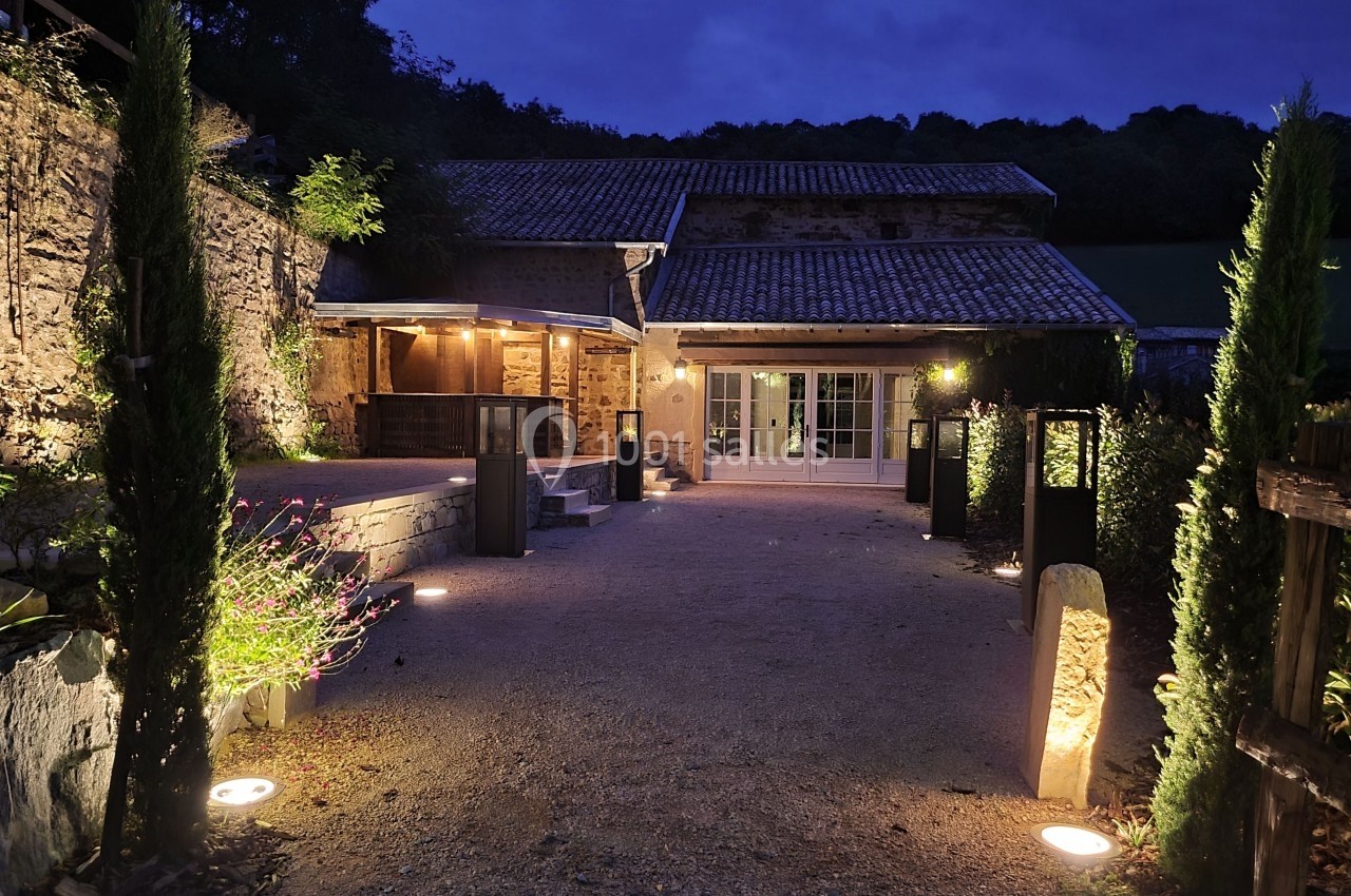 Cour extérieure éclairée d'une maison en pierre au crépuscule, entourée de végétation et de lampes au sol.