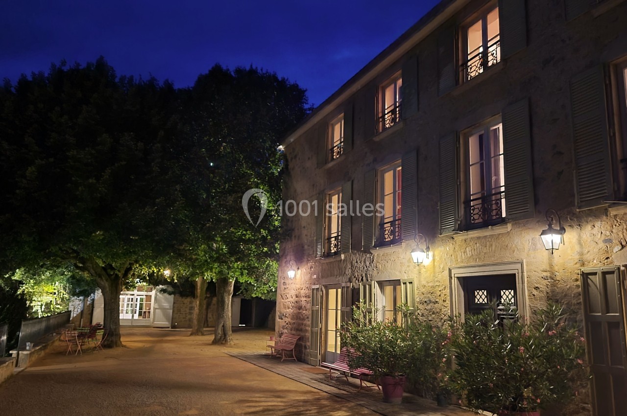 Façade éclairée d'une maison en pierre avec volets verts, arbres et bancs dans une cour au crépuscule.