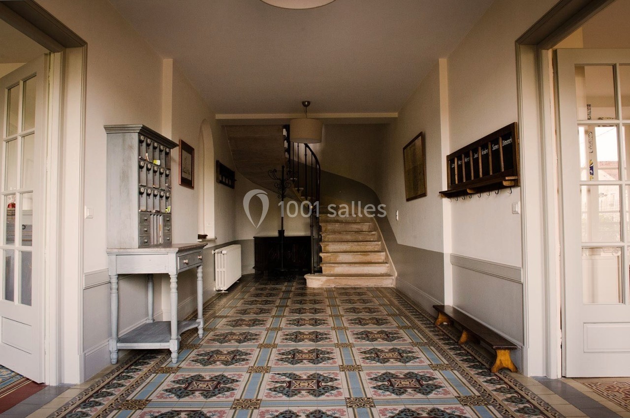 Couloir lumineux avec carrelage ancien, escalier en colimaçon au fond, meubles et portes vitrées sur les côtés.