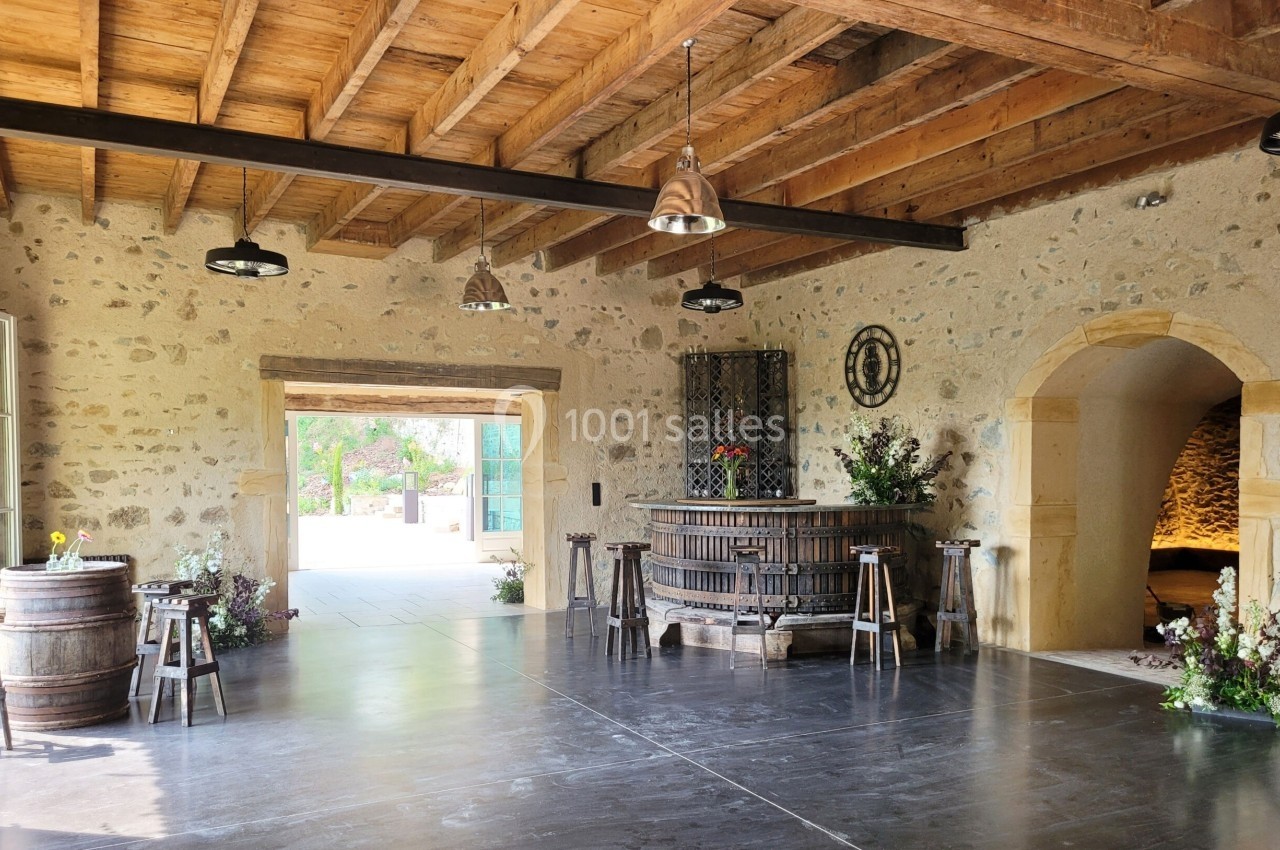 Salle en pierre avec un plafond en bois, un ancien pressoir au centre, des tonneaux et une ouverture sur l'extérieur.