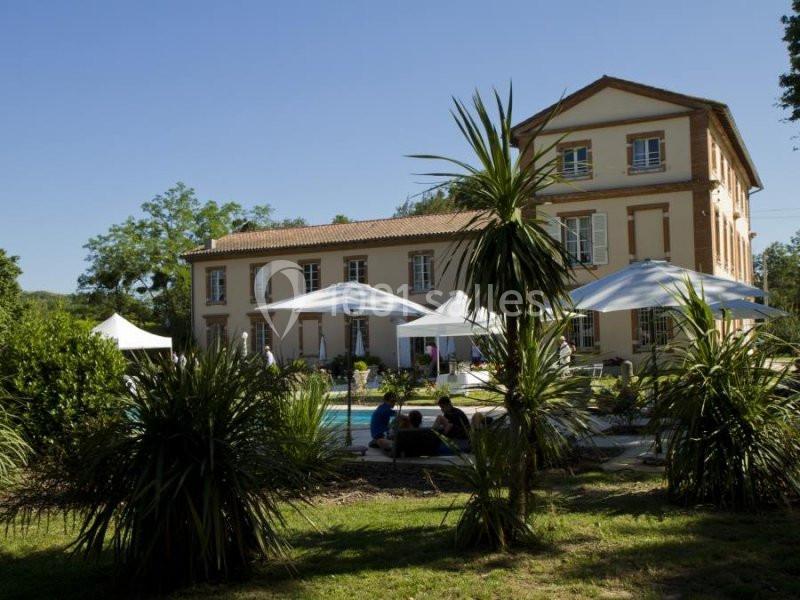 Grande maison à deux étages entourée de végétation, avec des parasols et des personnes près d'une piscine.