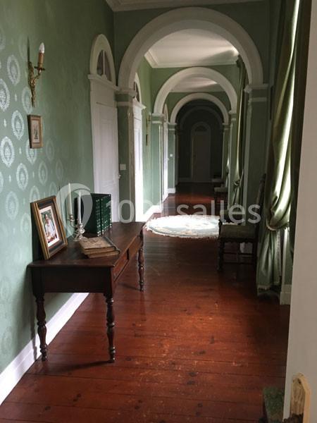 Couloir avec parquet en bois, murs verts décorés, table avec cadres et bougies, et rideaux verts aux fenêtres.