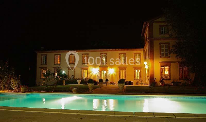 Façade éclairée d'une grande maison ancienne avec une piscine illuminée au premier plan, de nuit.