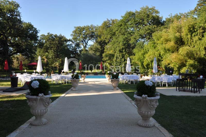 Allée bordée de plantes menant à une terrasse avec tables, chaises blanches et parasols, entourée de verdure.