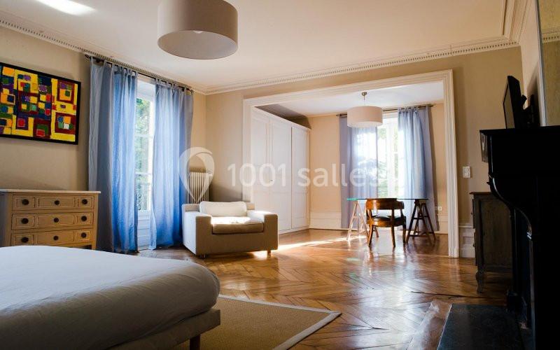 Chambre lumineuse avec parquet en bois, lit, fauteuil, table et grandes fenêtres avec rideaux bleus.