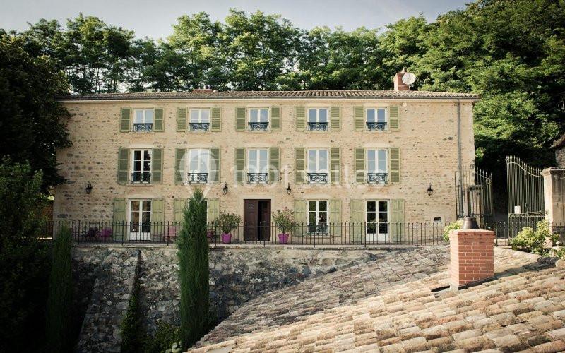 Façade d'une maison en pierre avec volets verts, entourée de végétation et située derrière une clôture en fer forgé.