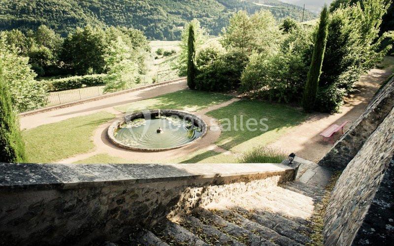 Vue d'un jardin avec une fontaine circulaire entourée de pelouse, bordé d'arbres et d'un chemin en pierre.
