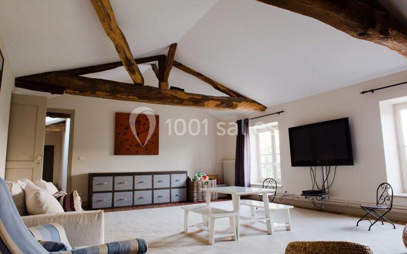 Salon lumineux avec poutres apparentes, mobilier moderne, grande télévision murale et fenêtres laissant entrer la lumière.