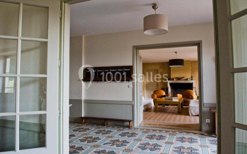 Vue intérieure d'une maison avec un sol en carreaux colorés, une pièce adjacente avec cheminée et fauteuils.