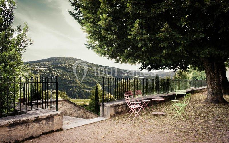 Chaises et table colorées disposées sous un arbre, avec vue sur une vallée verdoyante et des collines.