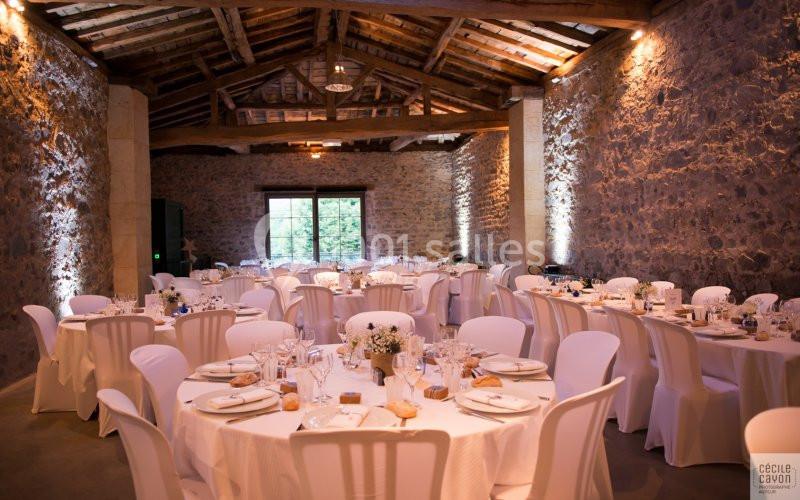 Salle en pierre avec plafond en bois, tables rondes dressées avec nappes blanches et lumière naturelle entrant par une…