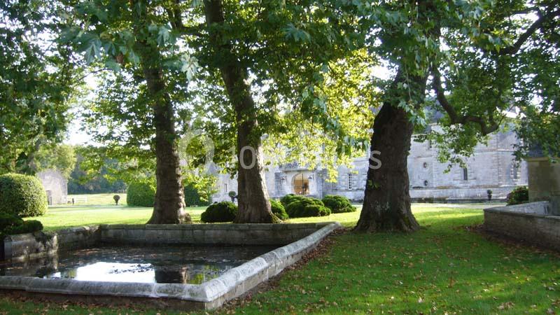 Vue d'un jardin avec un bassin rectangulaire entouré d'arbres et un bâtiment en pierre en arrière-plan.