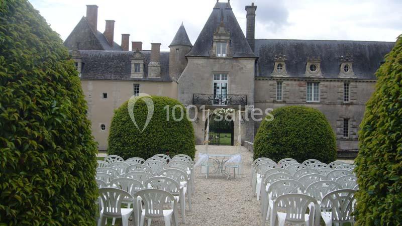 Allée centrale bordée de chaises blanches menant à une arche florale devant un château ancien en pierre.
