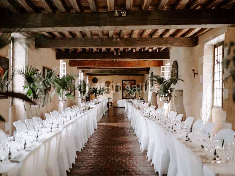 Salle de réception avec longues tables dressées, nappes blanches et décoration végétale sous un plafond en poutres…