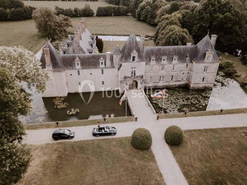 Vue aérienne d'un château entouré de douves, avec deux voitures garées devant l'entrée principale.