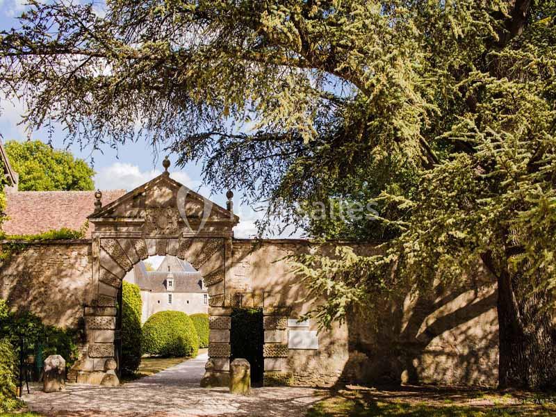 Entrée en pierre ornée d'une arche, donnant sur une cour avec un bâtiment ancien entouré de verdure.