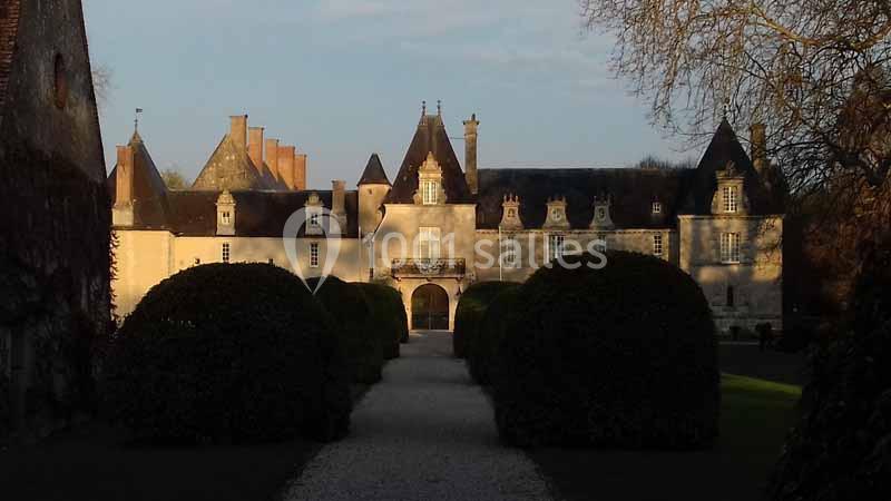 Façade d'un château en pierre avec des toits en ardoise, entouré de buissons taillés et d'une allée centrale.