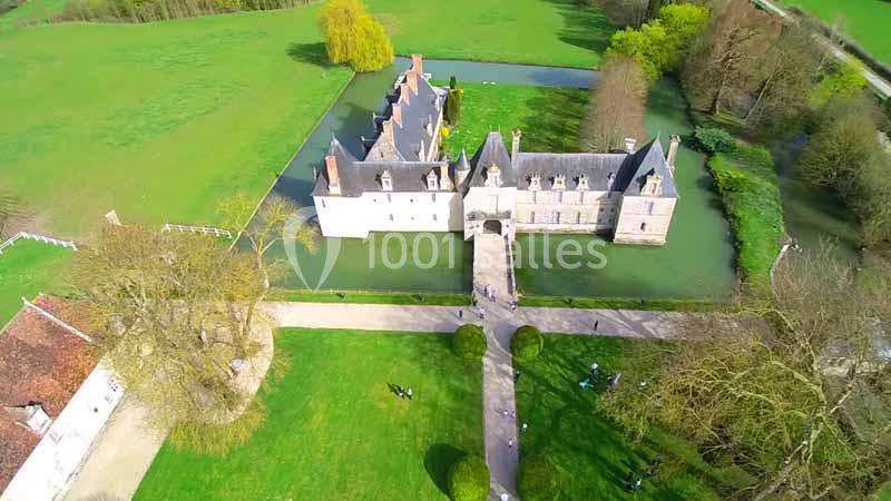 Vue aérienne d'un château entouré de douves, situé dans un paysage verdoyant avec des arbres et des pelouses.