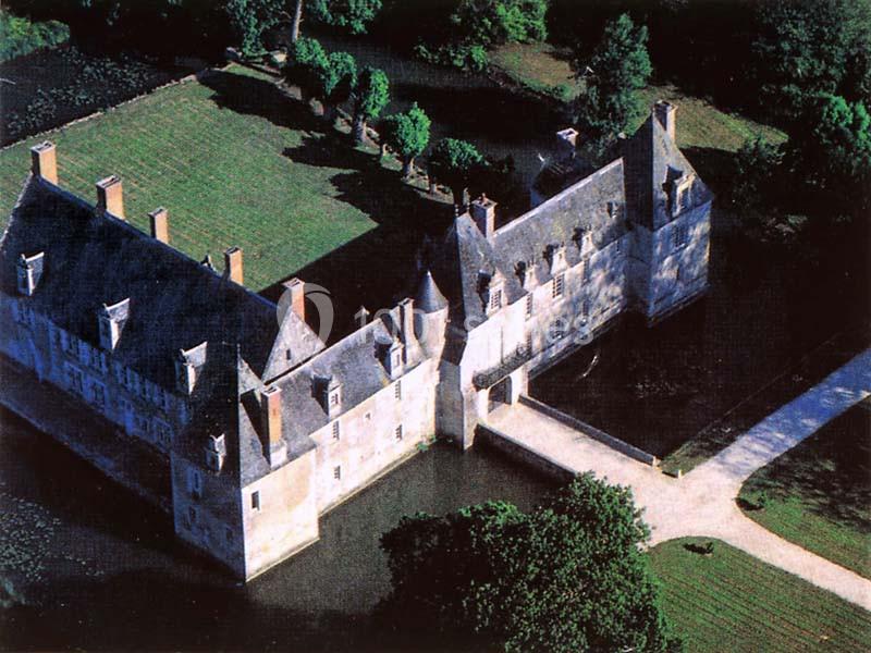 Vue aérienne d'un château entouré d'eau, avec un pont menant à l'entrée principale et des arbres en arrière-plan.