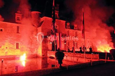 Façade d'un château illuminée par des lumières rouges, avec des drapeaux et des spectateurs en arrière-plan.