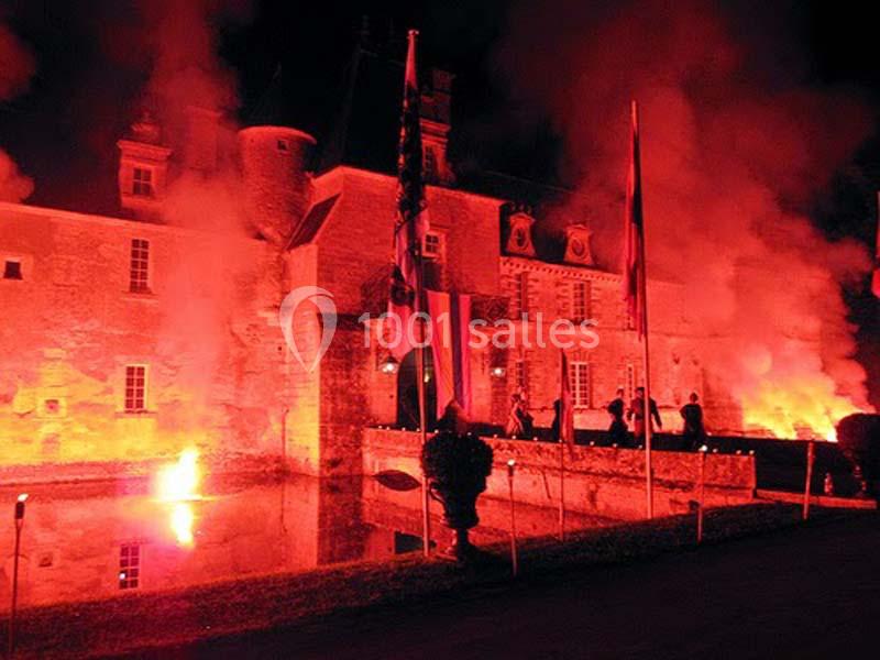Façade d'un château illuminée par des lumières rouges, avec des drapeaux et des spectateurs en arrière-plan.