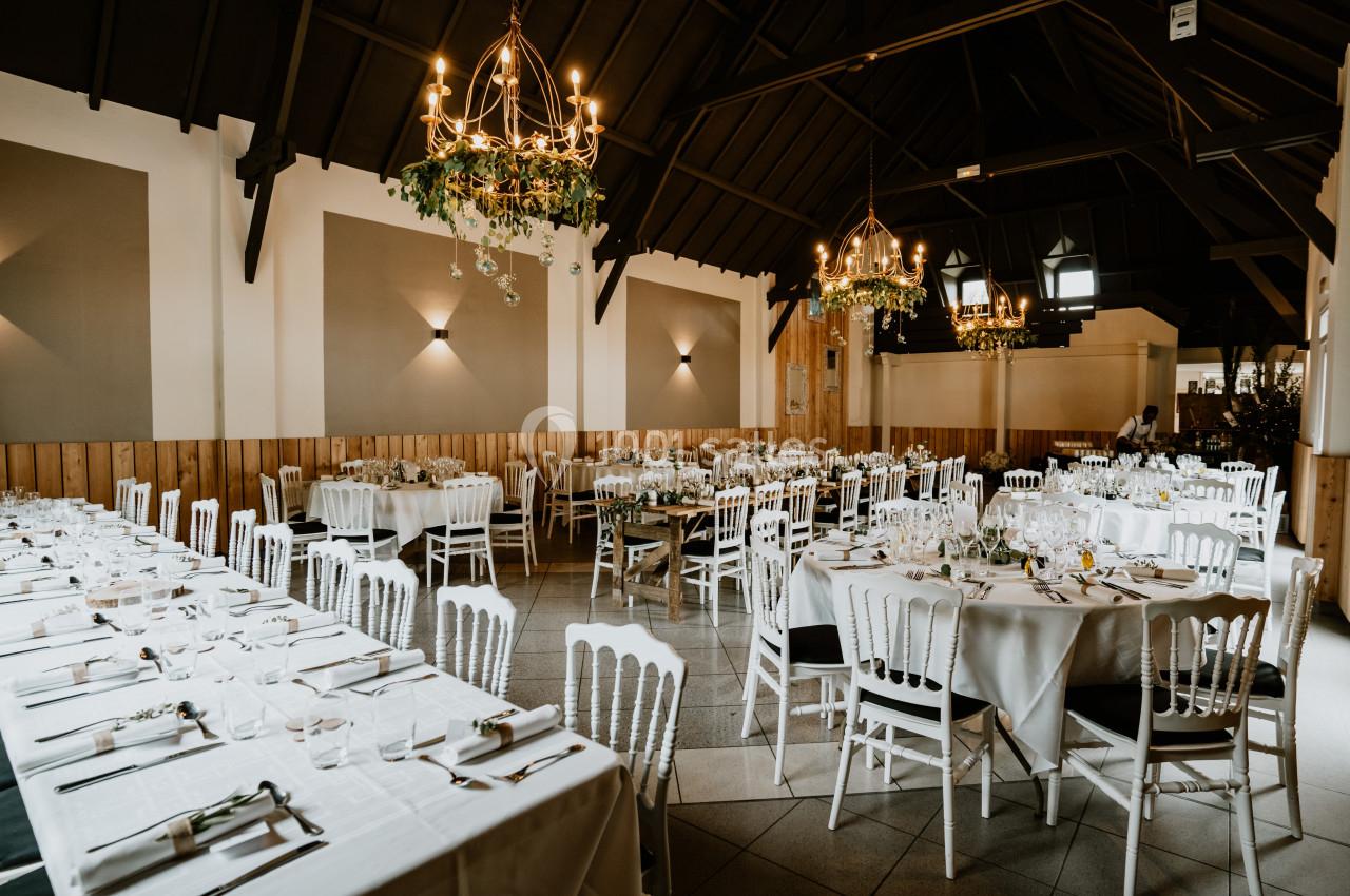 Salle de réception élégante avec tables dressées, chaises blanches et lustres suspendus dans un décor chaleureux.