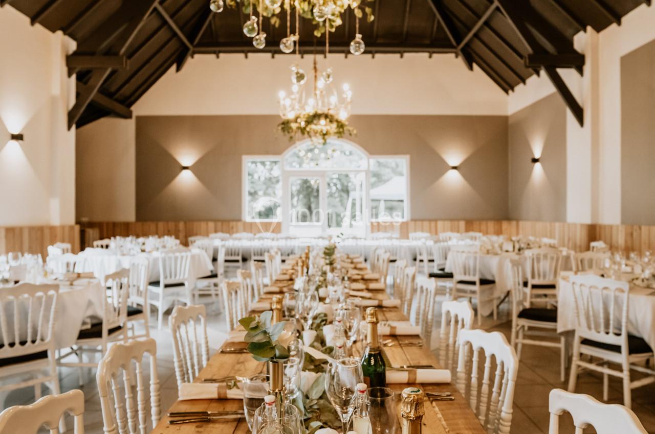 Salle de réception décorée avec des tables en bois, chandeliers suspendus et chaises blanches alignées.