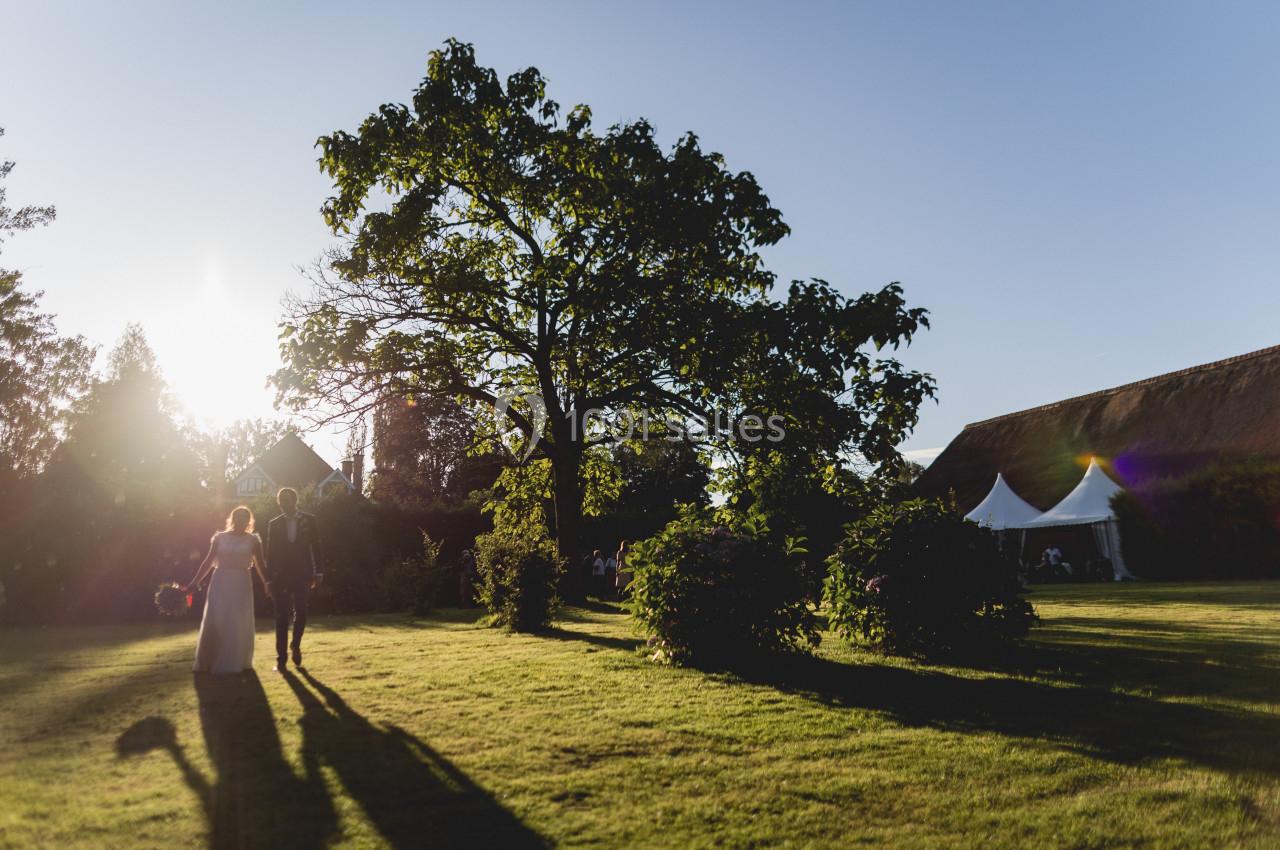 Un couple marche sur une pelouse au coucher du soleil, près d'un grand arbre et de tentes blanches.
