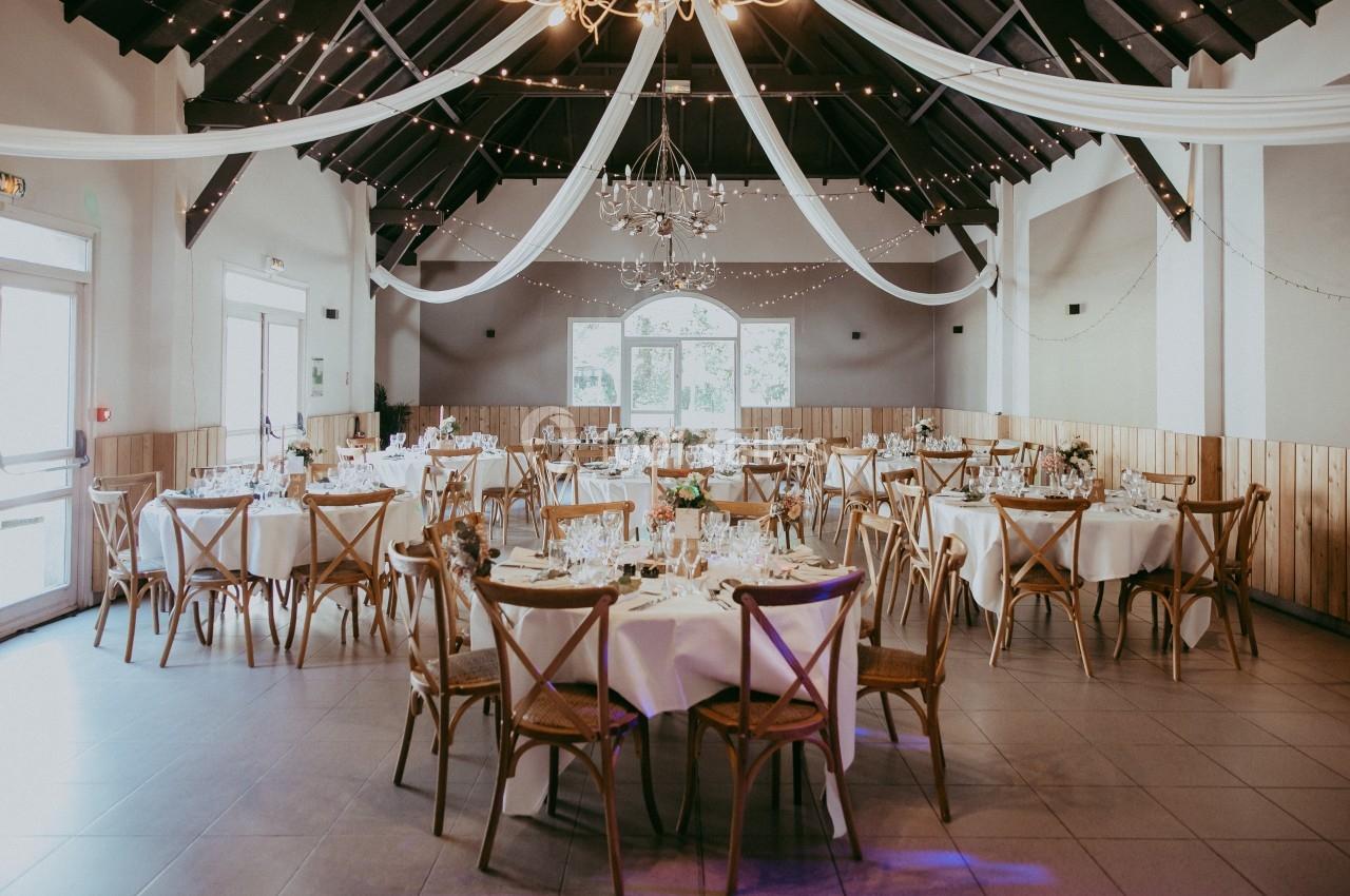 Salle de réception décorée avec des tables rondes dressées, des chaises en bois et des guirlandes lumineuses suspendues.
